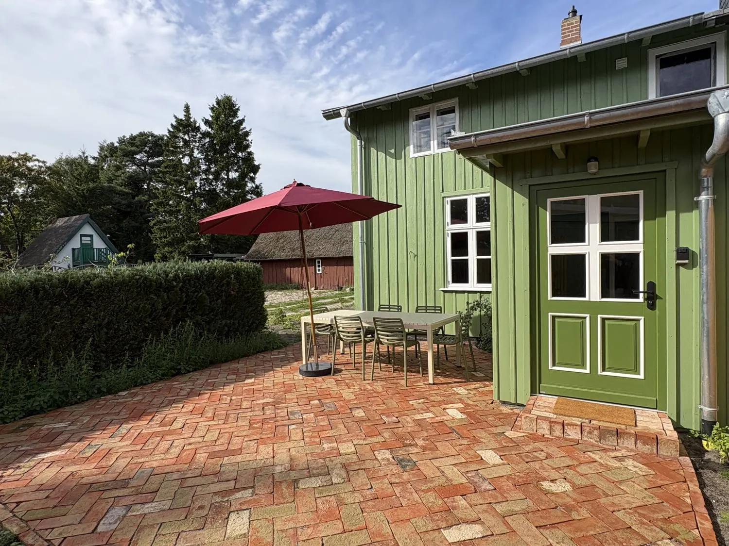 Ferienhaus Prerow: Terrasse mit Klinkerpflaster, Esstisch, Stühlen und rotem Sonnenschirm vor der grünen Haustür