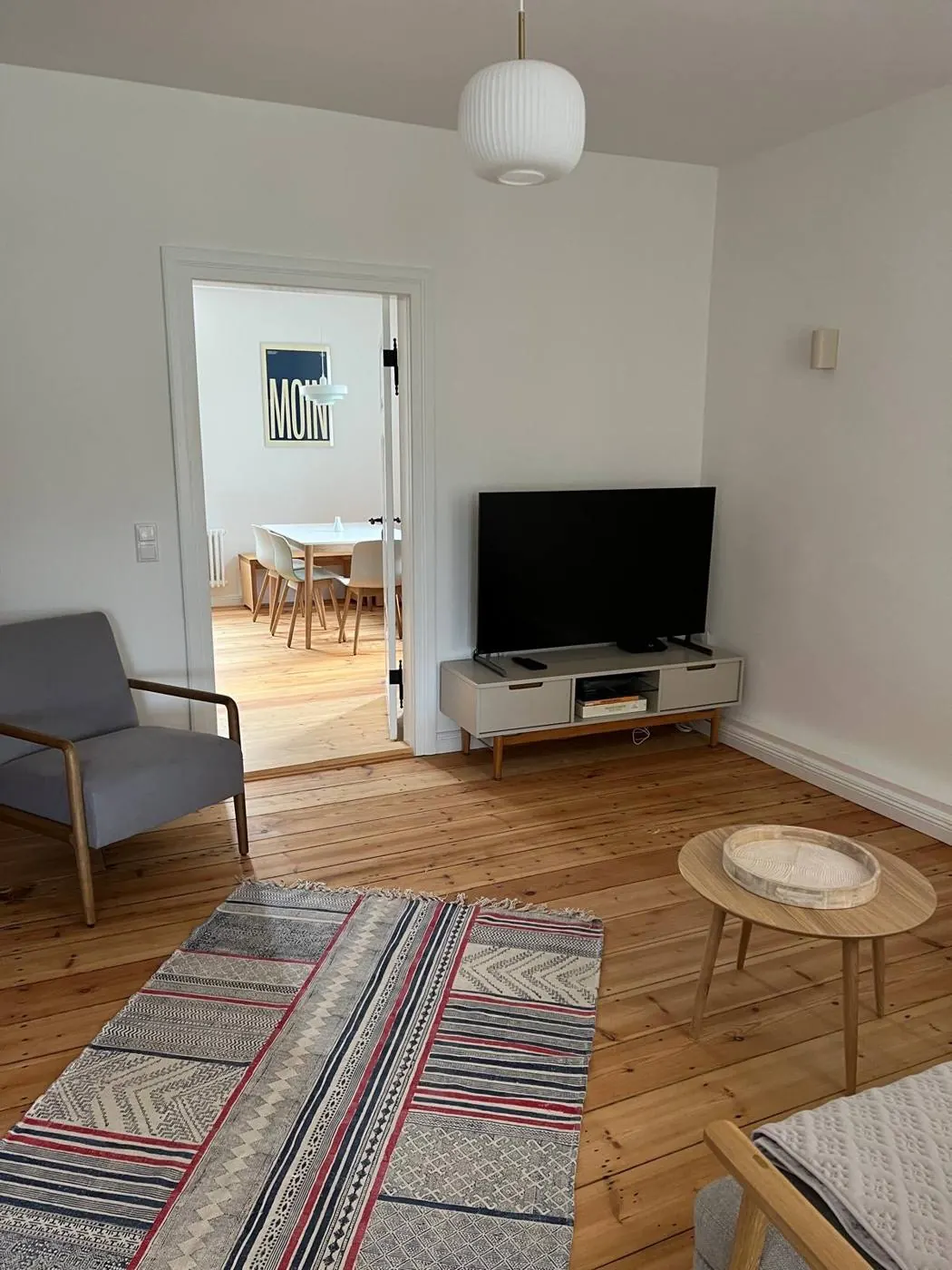Wohnzimmer mit Fernseher, Sessel und gemustertem Teppich, im Hintergrund die Küche